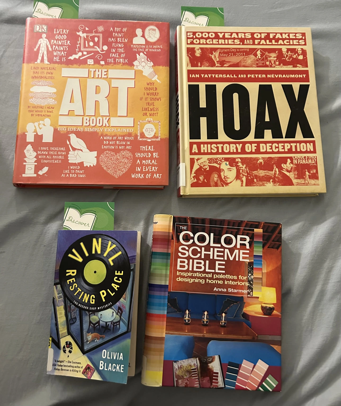 An image of four books sitting on a bed. From top left to bottom right: 'The Art Book: Big Ideas Simply Explained', 'Hoax: A History of Deception' by Ian Tattersal and Peter Nevraumont, 'Vinyl Resting Place' by Olivia Blacke, and 'The Color Scheme Bible: Inspirational Palettes For Designing Home Interiors' by Anna Starmer.